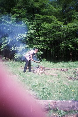 A man in 1970s attire tends to a small campfire in a lush, grassy clearing framed by dense, green trees. Smoke rises visibly....