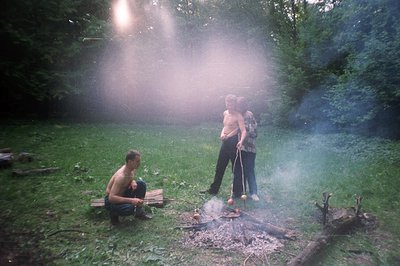 A candid moment captured in a wooded area; two young men are roasting food on skewers over an open fire. The scene is envelop...