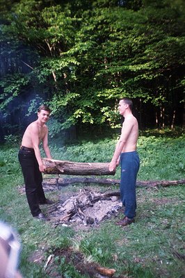 Two shirtless men carry a large log near a small, smoldering campfire. Set within a dense, green woodland environment. Appear...