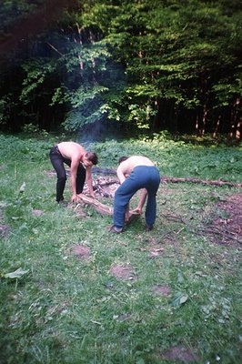 Two men, one shirtless, are tending a small campfire in a lush, grassy clearing within a dense forest. Both wear relaxed-fit,...