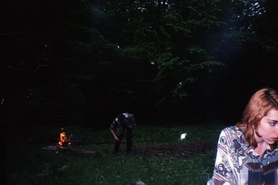 A nighttime outdoor scene: a small campfire burns in a grassy area framed by dense foliage. Two figures are present - one ten...