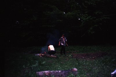 A bonfire illuminates two figures in a wooded area. A woman kneels tending the fire, while a man stands nearby wearing a shor...