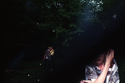 A grainy, partially obscured portrait shows a woman partially obscuring the lens with her hand. Two figures stand in the back...