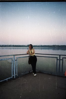 A woman stands on a concrete overlook, gazing towards a wide body of water at dusk. She wears a yellow top, black pants, and ...