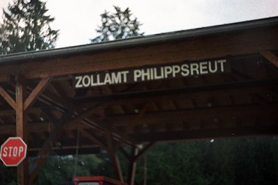 A weathered wooden sign reads "ZOLLAMT PHILIPPSREUT" against a backdrop of dark, forested foliage. A red stop sign is partial...