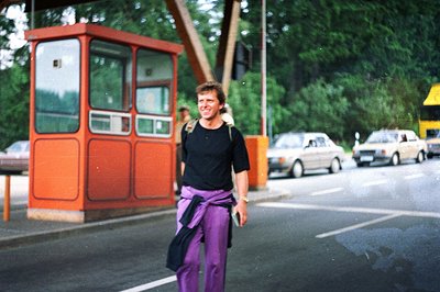 A man stands beside a roadside control booth, a weathered orange structure, circa 1970s. He wears a black shirt, purple pants...