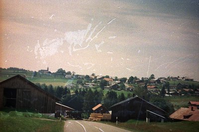 Rural landscape view featuring timber farmhouses and steeply sloped roofs nestled within a valley. Visible alpine peaks domin...