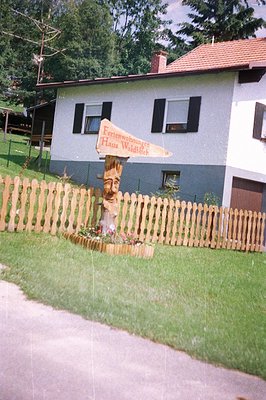 A two-story house with a steeply pitched roof stands on a slope, displaying a sign reading "Ferienwohnungen Haus Waldblick." ...