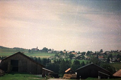 Rural landscape featuring wooden barns and a distant village, captured on film. Visible wear and grain characteristic of olde...