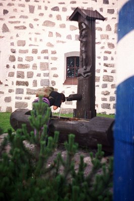 A weathered, intricately carved wooden statue stands atop a stone base, set within a courtyard. A person in a hooded sweatshi...