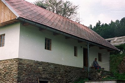 Stone-clad base supports a white-washed, steeply-roofed building with a small window. A man sits on a small, raised porch. Li...