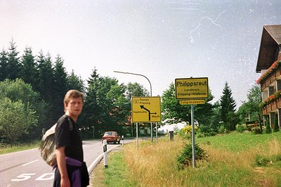 A young man stands beside a road sign in Philippsreut, Germany. The sign directs to Freying and Grafenau, noting a customs bo...