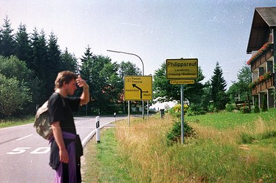 A solitary traveler in purple separates stands by a roadside sign for Philippsreut, near Freyung, Germany. The scene suggests...