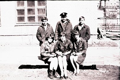 Six teenagers pose outside a brick building with a visible industrial tank in the background. Three males stand, two with sho...