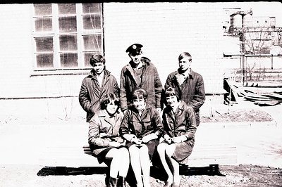 Group portrait of six young people, likely students, outdoors. Three are seated on a wooden bench, three stand behind. The in...