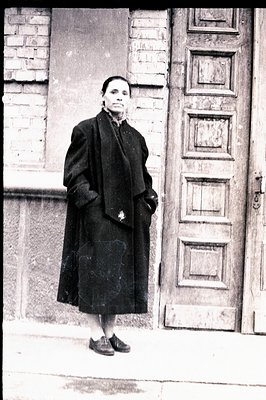 Full-length portrait of a woman in a long, dark coat, standing before a paneled wooden door and brick wall. The image exhibit...