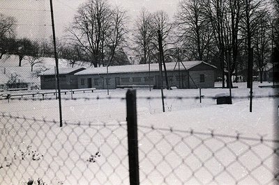 A long, single-story building with a gently sloped roof sits amidst a snow-covered landscape. Bare trees frame the structure,...
