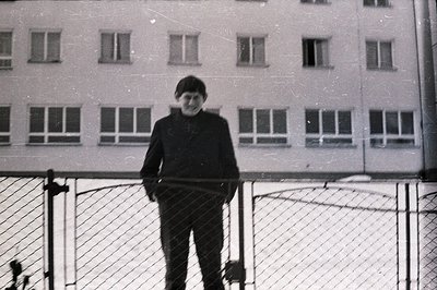 A young man stands near a chain-link fence, positioned against a stark, multi-windowed building. He wears a dark suit jacket ...