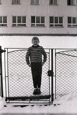 A young boy stands between a chain-link fence, partially obscured, in a snowy landscape. He wears a patterned sweater, dark p...