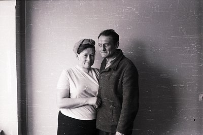A formally posed portrait of a couple, likely taken indoors against a plain wall. The woman wears a beret and a simple white ...