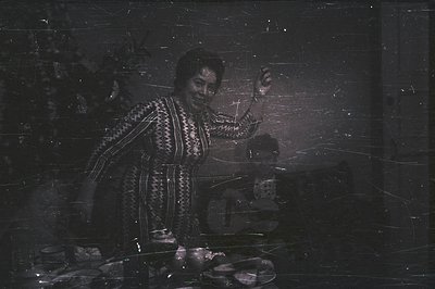 A warmly lit scene depicts a woman in elaborate, patterned dress gesturing expressively with raised arms. A blurry figure sta...