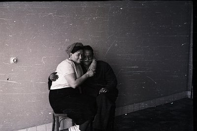 A pair embrace in a stark, intimate portrait. The man wears a cap and dark trousers; the woman, a light-colored top. A single...