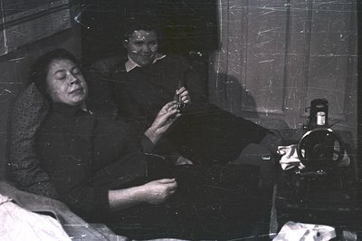 A woman sits and appears to be sewing, with a second woman smiling slightly behind her. A vintage camera sits on a nearby tab...