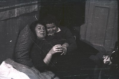 Two figures, an older woman and younger man, are closely positioned on a patterned sofa. The woman is gazing intently, while ...