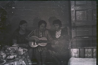 Three individuals are seated; a guitarist center, flanked by two others. The scene appears to be a domestic interior, with vi...