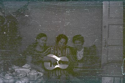 Three men with long hair are seated and holding guitars in what appears to be a domestic interior. The photo exhibits signifi...