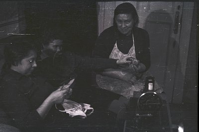 Two women sit closely, meticulously repairing shoes within a dimly lit interior, likely a workshop or home. Visible tools and...