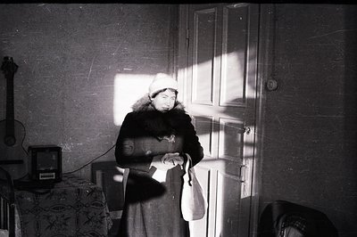 A woman in a fur coat and headscarf stands in a doorway, dramatically lit by sunlight. A guitar rests on the wall, and a patt...