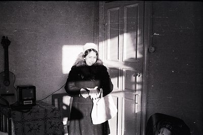 A woman in a dark coat and head covering stands framed by a doorway, light dramatically highlighting her face and silhouette....