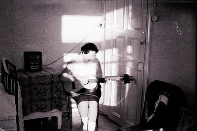 A woman seated, playing a guitar in a sunlit room. The strong light source, visible through a paneled door, casts dramatic sh...