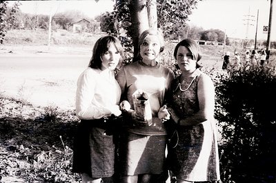 Three individuals stand outdoors; two young women flank a mature woman holding a cylindrical object. The scene appears to be ...