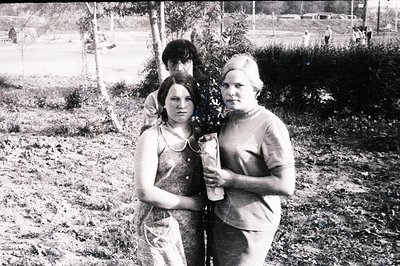 Three young individuals stand in a grassy, open field, likely a park or recreational area. The two women are prominently feat...