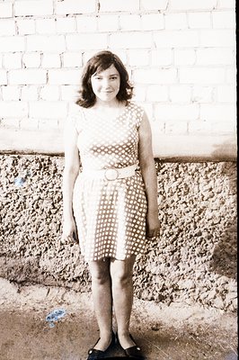 A young woman poses outdoors in a vintage dress with a white belt. She wears black flats. The background features textured st...