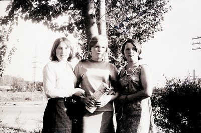 Three young women stand outdoors, posing near a large tree. They wear 1970s-era dresses. One holds a small package or book. A...