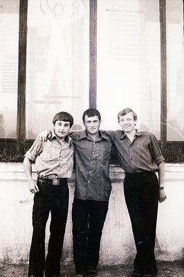 Three young men, dressed in 1970s-era casual attire, stand close together, arms around each other's shoulders, posing in fron...