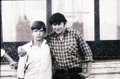 Two young men stand close together in a posed portrait. One wears a button-down shirt, the other a plaid flannel over a white...