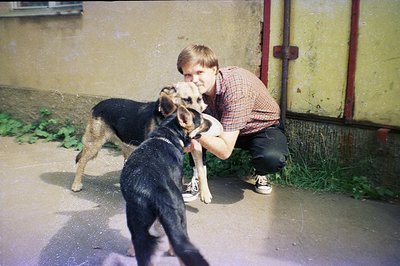 A young man crouches, embracing a medium-sized, dark-furred dog with tan markings. The backdrop shows a weathered, textured w...
