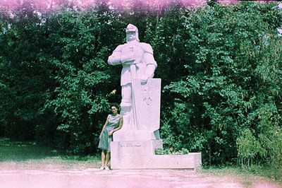 A woman in a short-sleeved dress poses alongside a monumental, stylized sculpture of a soldier, likely commemorating military...