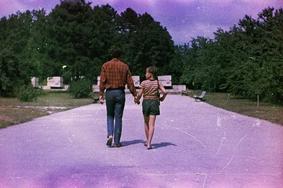A man and a young child walk hand-in-hand along a paved path, receding into a wooded area. The man wears jeans and a red plai...
