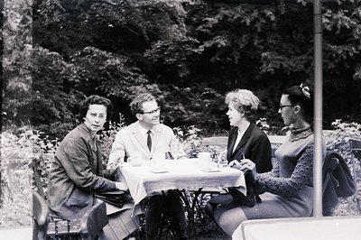 Four people are seated around a small, fabric-covered table in a garden setting. The men wear suits, while the women sport st...