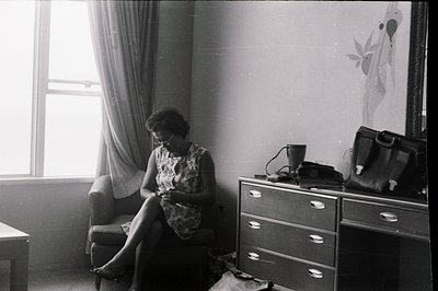 A seated woman, appears to be knitting in a well-lit room. Mid-century modern furniture & wallpaper indicate a likely 1960s s...