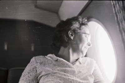 A middle-aged woman in a patterned top gazes out a circular window, likely on a train or ship. Interior details suggest a mid...