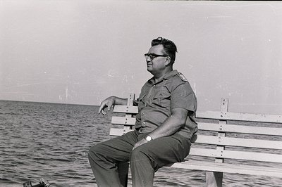 A man in a military-style uniform sits on a wooden bench overlooking the sea. He wears glasses and a wristwatch. The image, l...