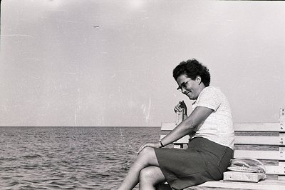 A seated woman with a short, curly hairstyle and glasses rests on a wooden bench overlooking the sea. She wears a short-sleev...