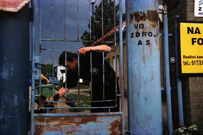 A man wearing a dark t-shirt is visible peering through a rusted, blue metal gate. The text "VOD ZDROJ A.S." is printed on th...