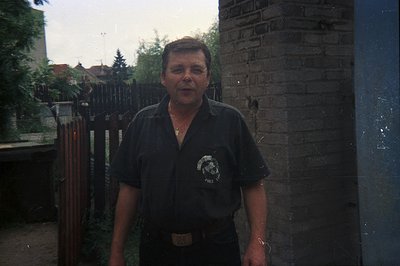 A man stands outdoors in front of a brick wall with visible signs of age and minor damage. He's wearing a dark short-sleeved ...